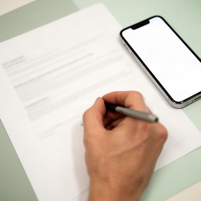 Hand schreibt eine formelle Anfrage auf Papier neben einem Smartphone